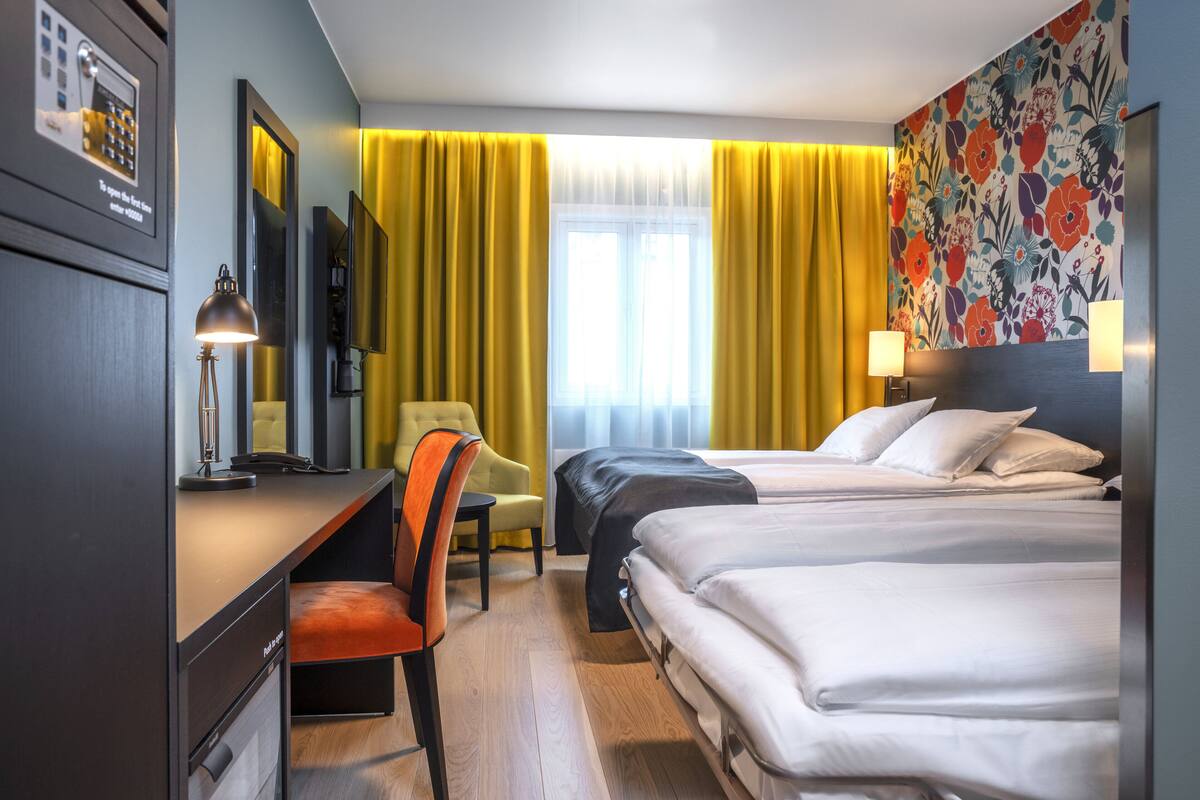 A hotel room with two beds, a desk, a chair, a desk lamp, and a large window with curtains.