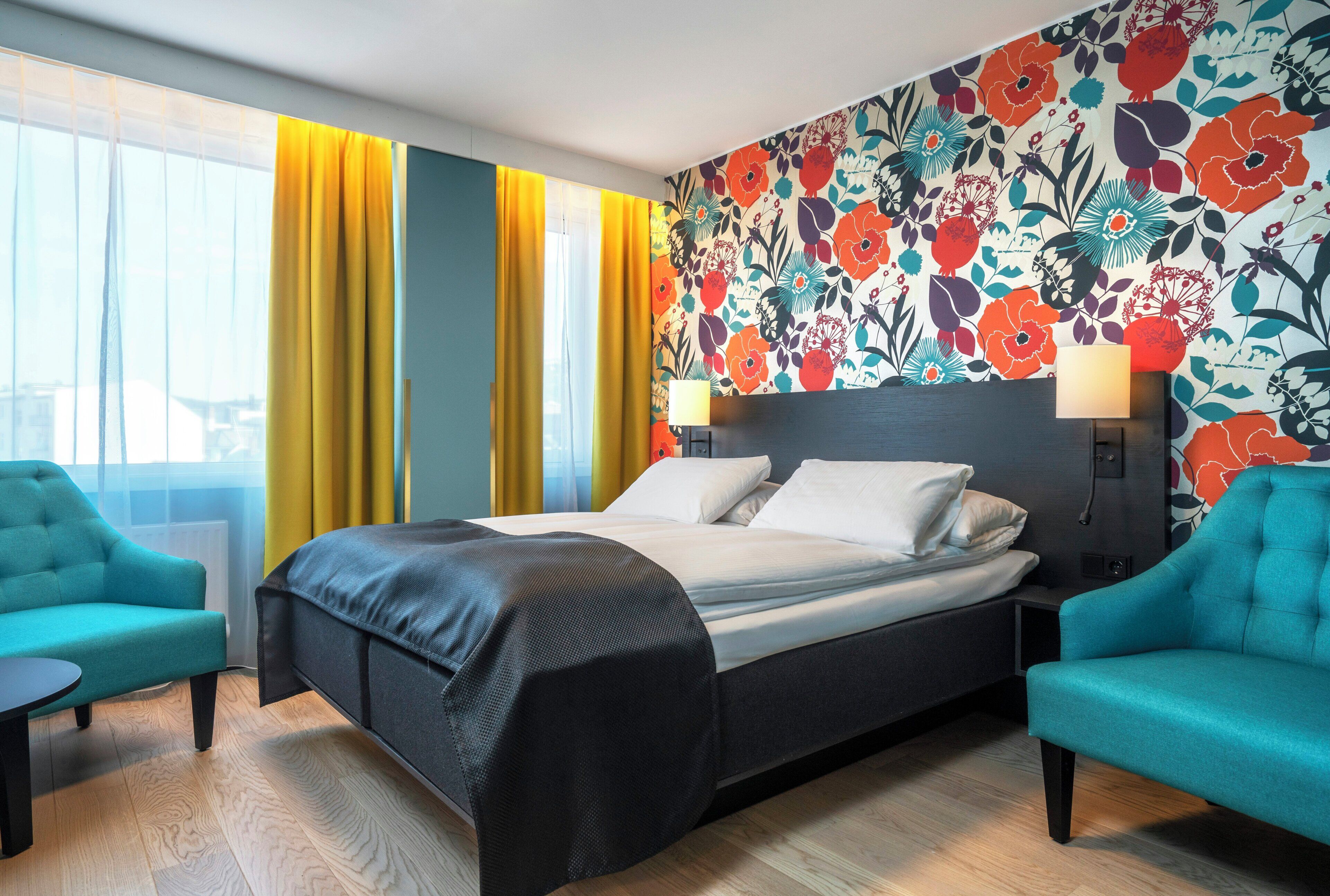 Habitación de hotel con cama, dos sillas, una mesita de noche y papel tapiz con motivos florales.