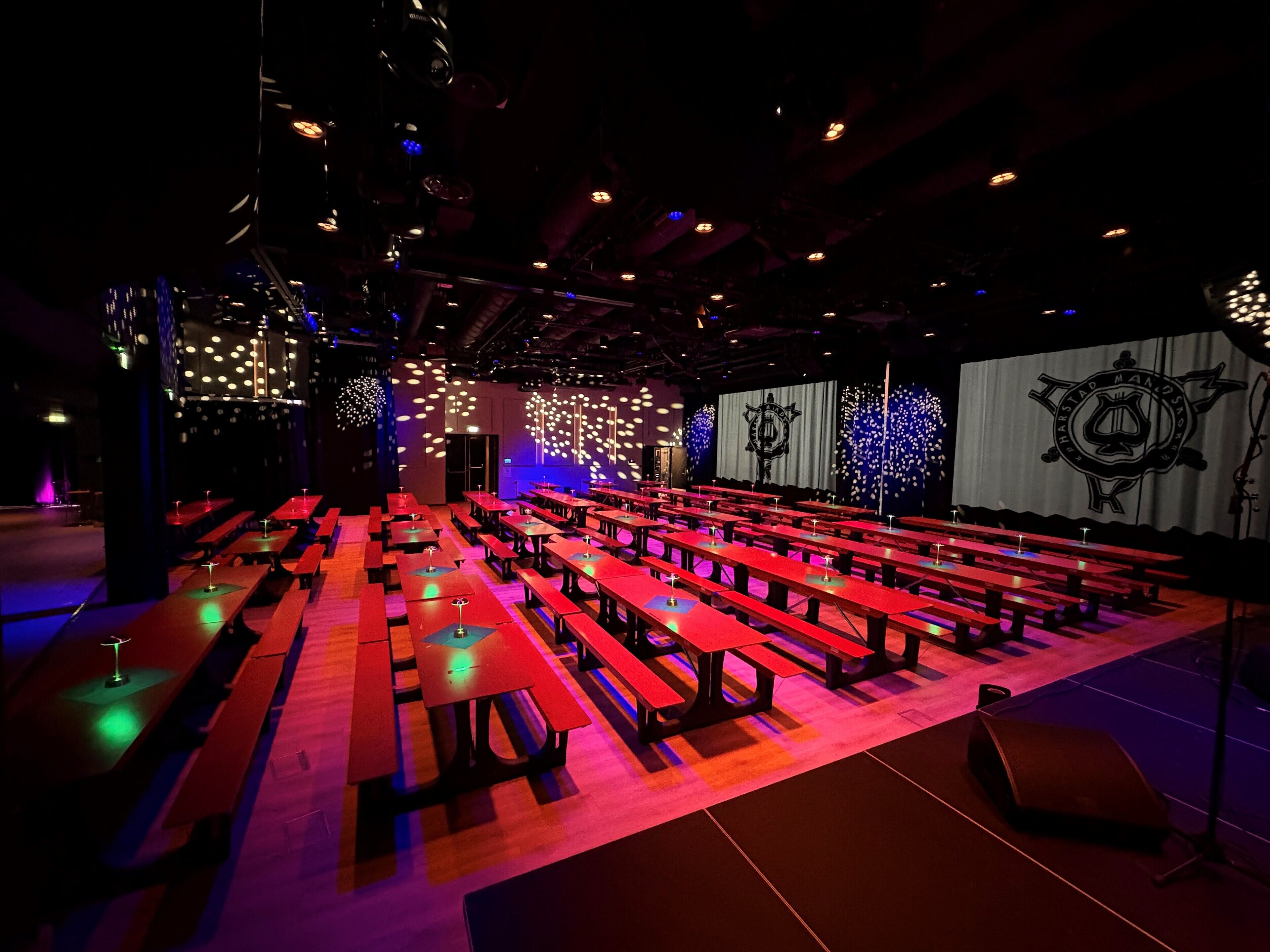 An indoor venue with rows of red tables and benches, a stage with a backdrop, and a ceiling with hanging lights.