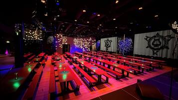 An indoor venue with rows of red tables and benches, a stage with a backdrop, and a ceiling with hanging lights.