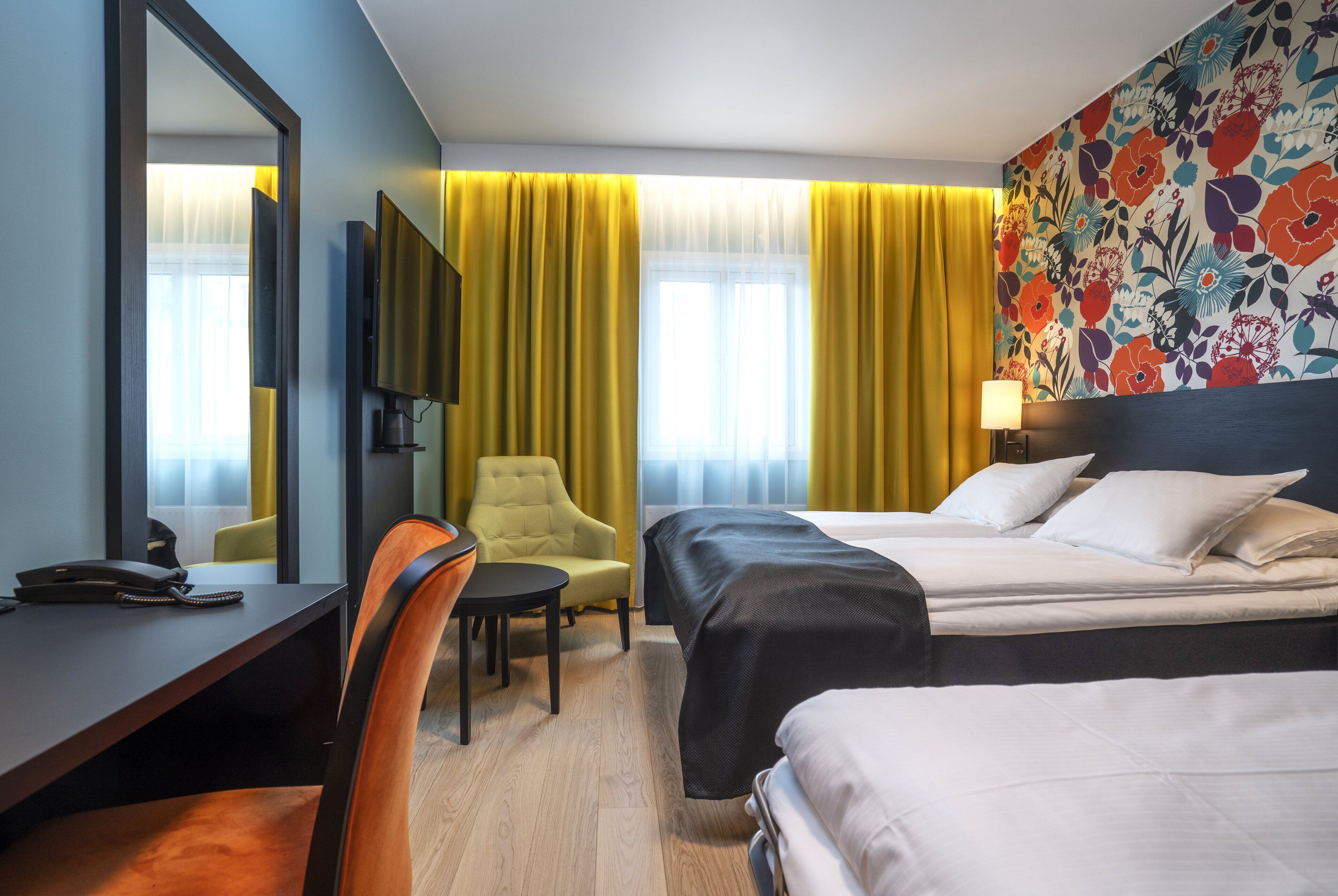 A hotel room with a desk, two beds, a chair, a TV, and a large window with curtains.
