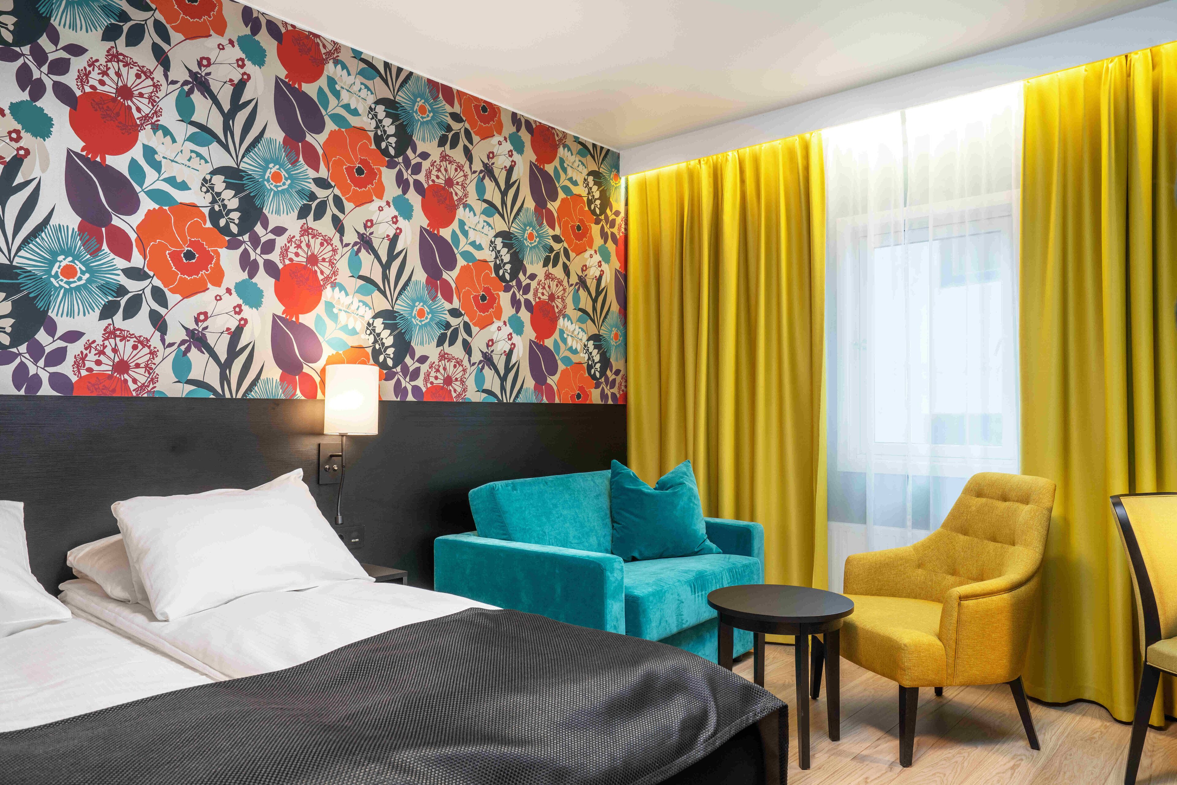 A hotel room with a bed, a teal sofa, a yellow armchair, and a floral-patterned wall.