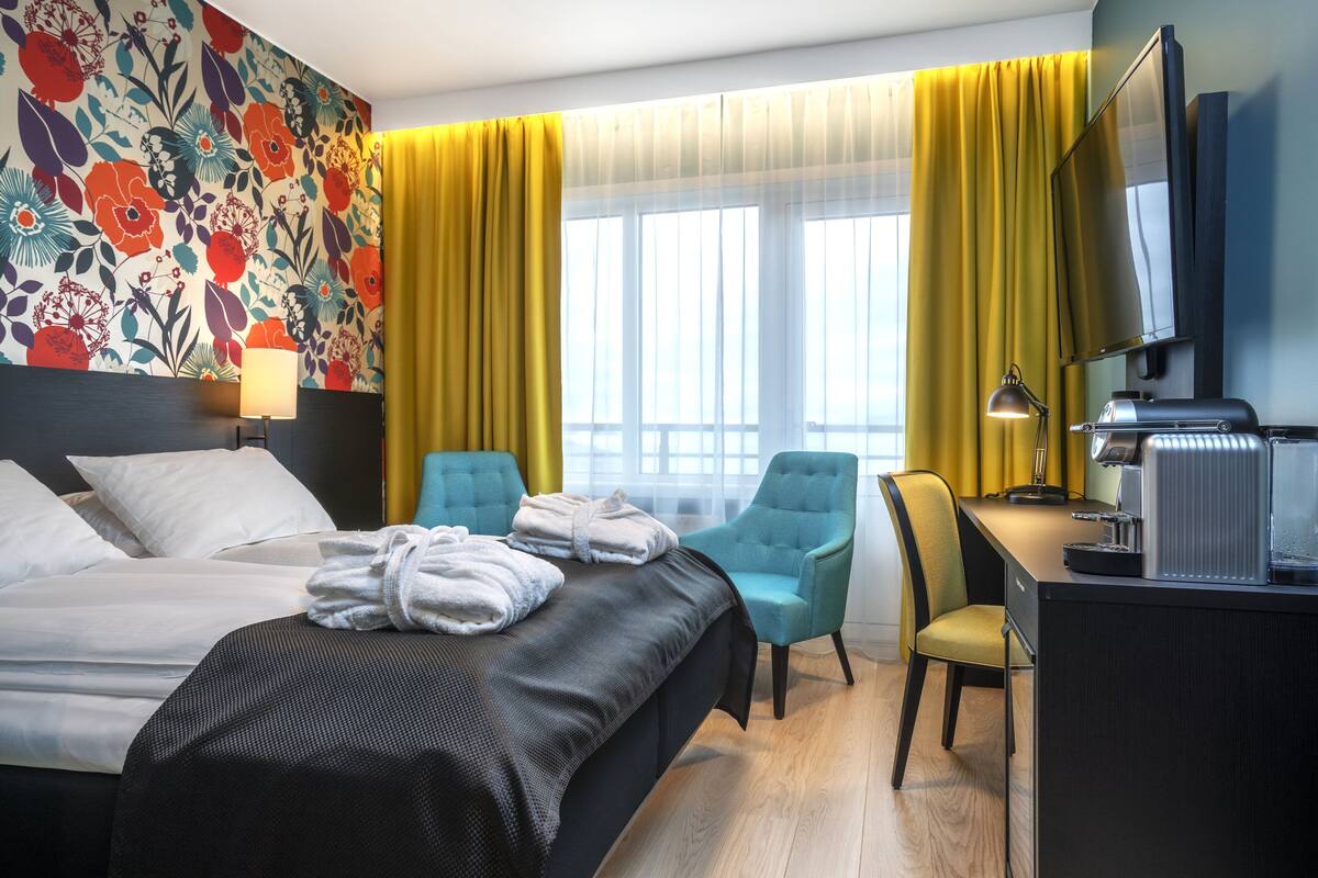 A hotel room with a bed, two chairs, a desk with a computer, a TV, and a window with yellow curtains.