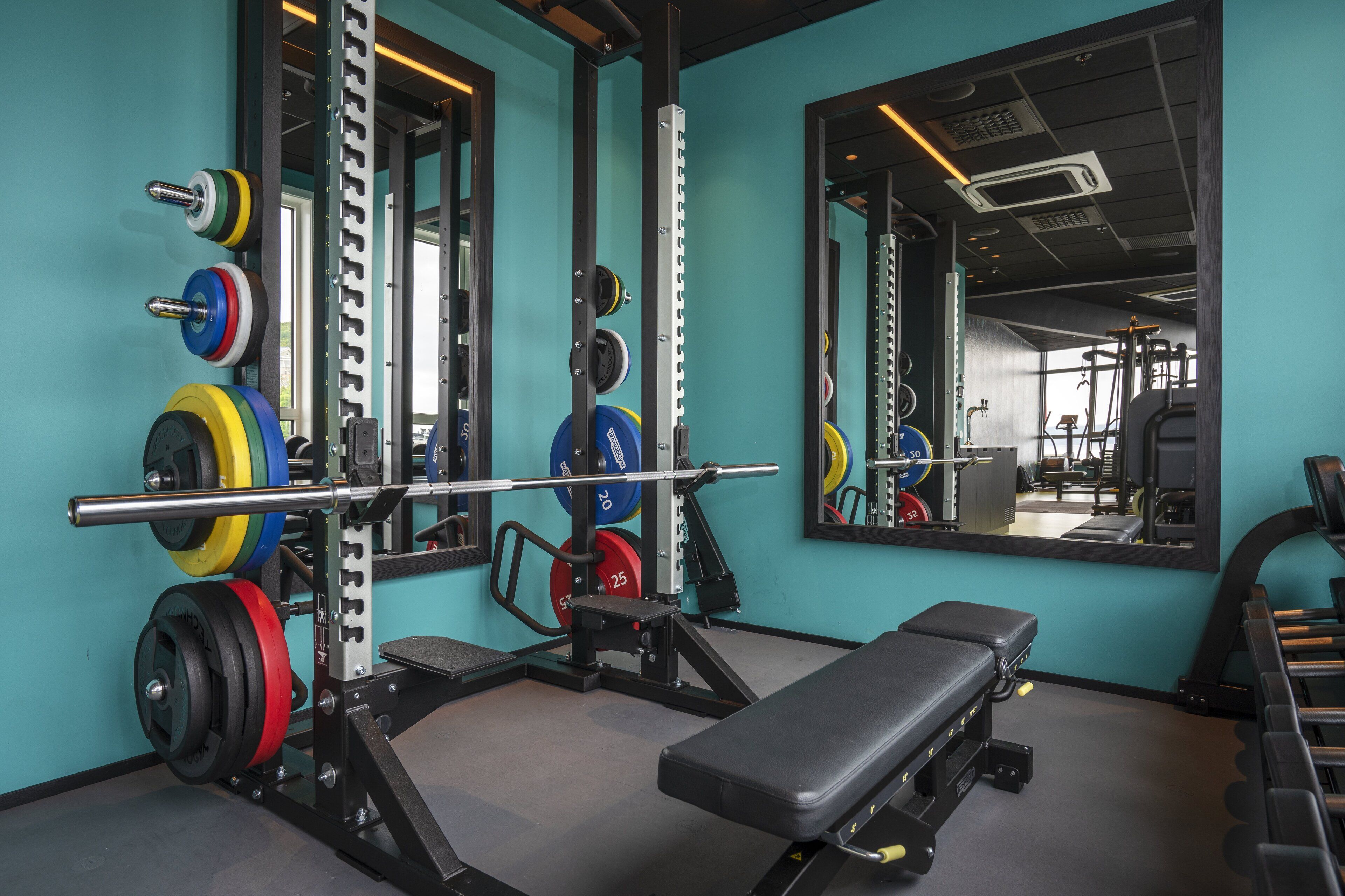 A gym with weightlifting equipment, a bench, and a mirror reflecting the equipment.