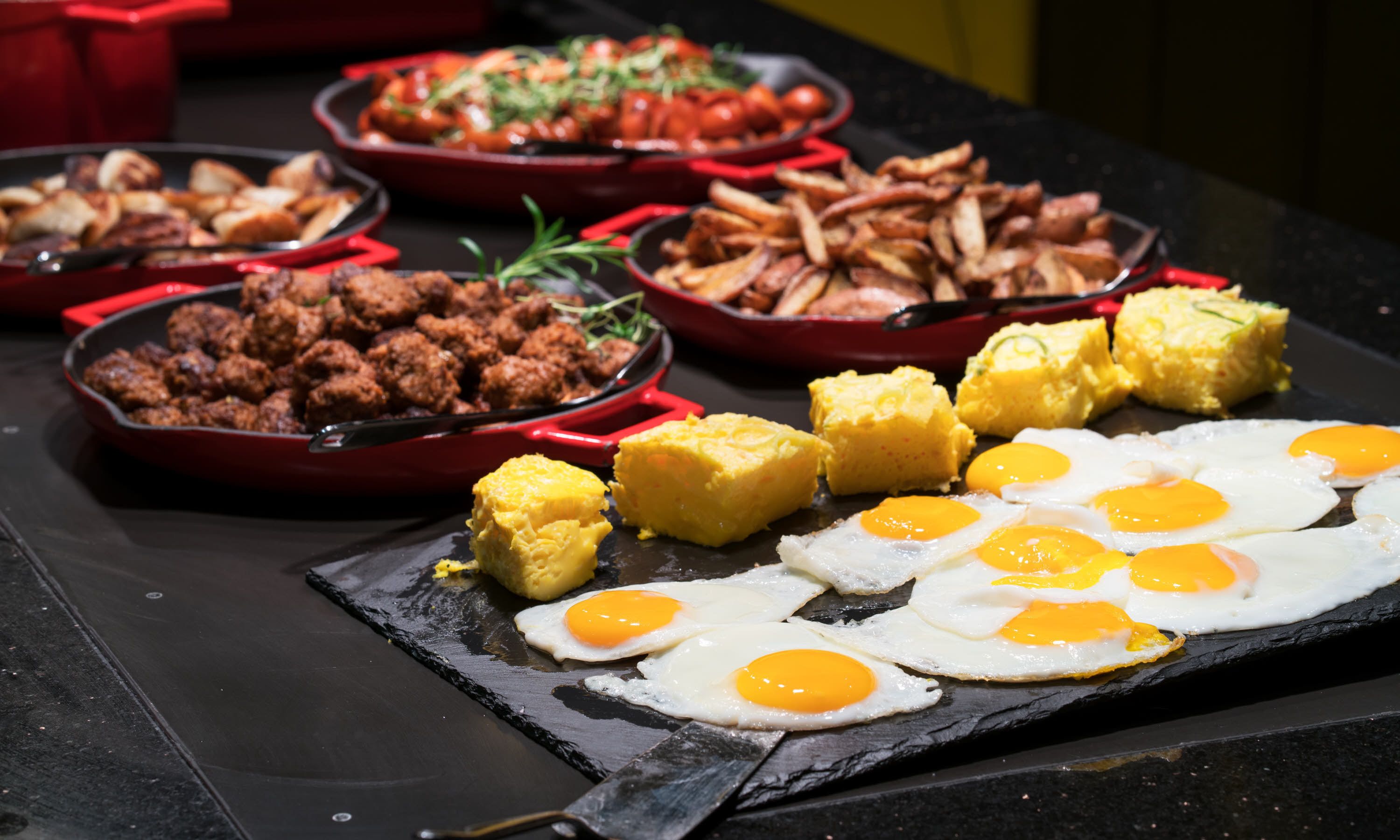 Buffet de café da manhã com diversos pratos, incluindo ovos mexidos, ovos fritos e diferentes tipos de carnes e vegetais.