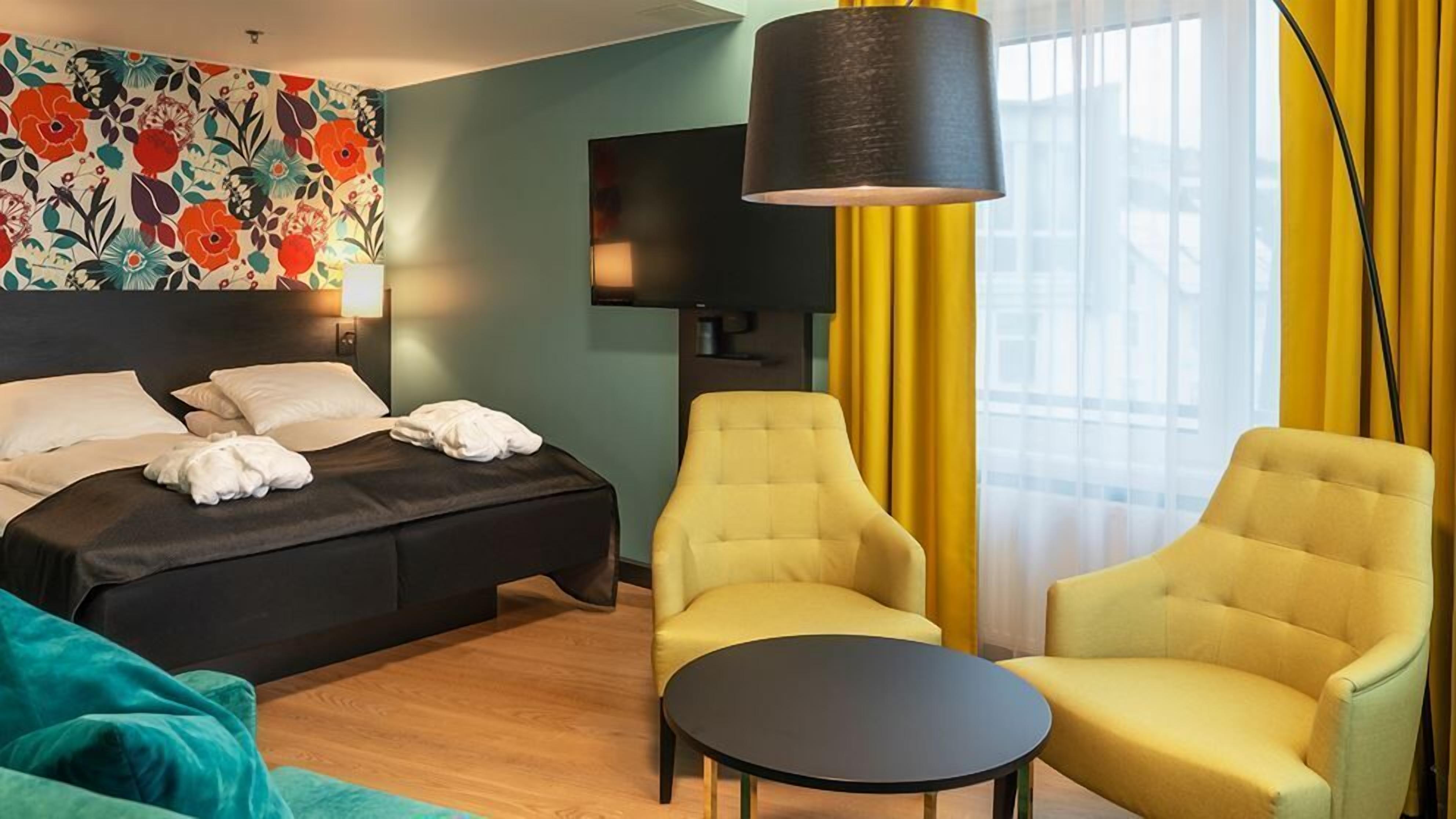 A hotel room with a bed, two armchairs, a small table, a TV, and a floral wall decoration.
