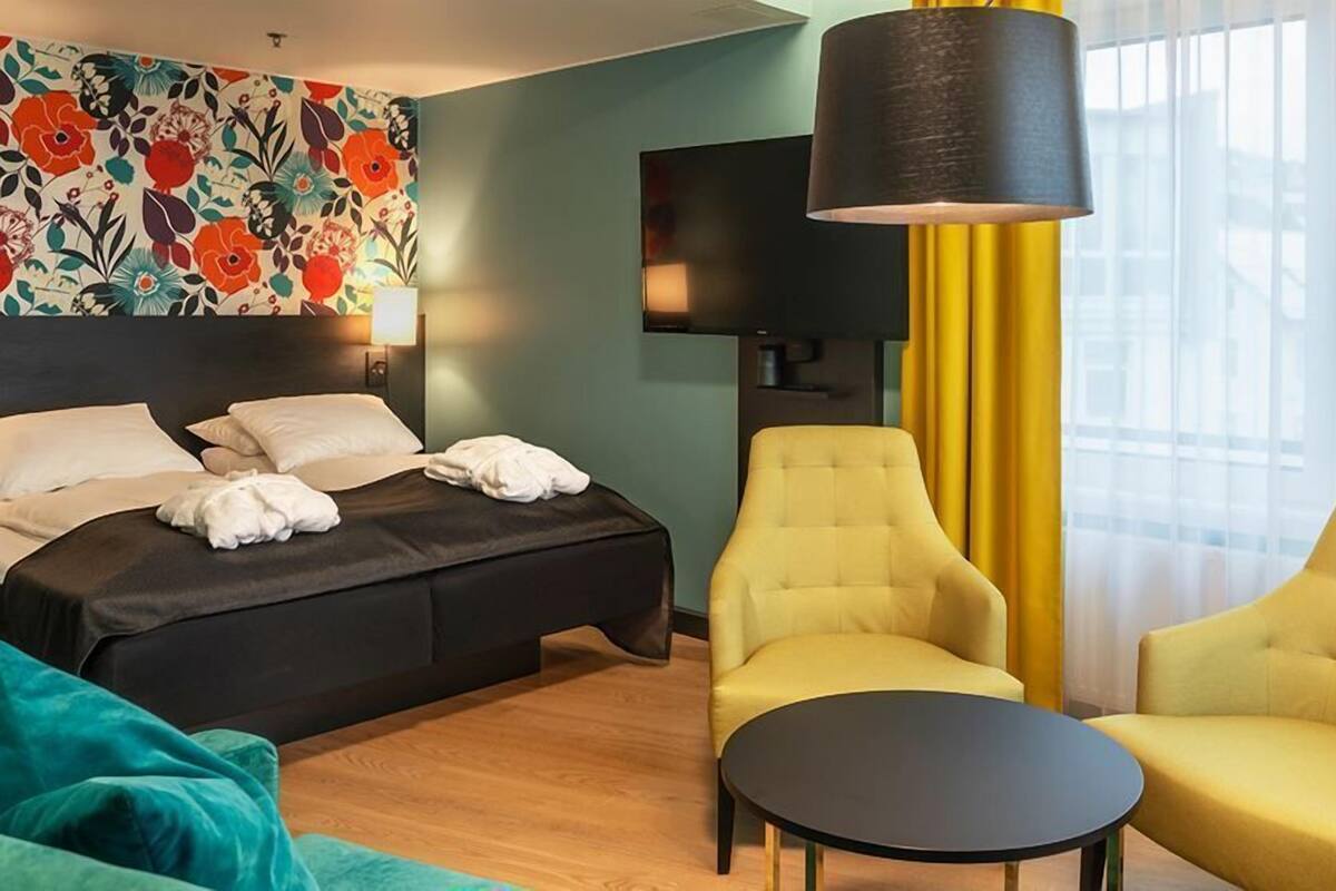 A hotel room with a bed, two armchairs, a small table, a TV, and a floral wall decoration.
