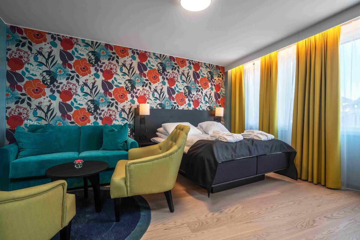 A hotel room with a bed, a green sofa, a yellow armchair, a small table with a vase, and a floral-patterned wall.
