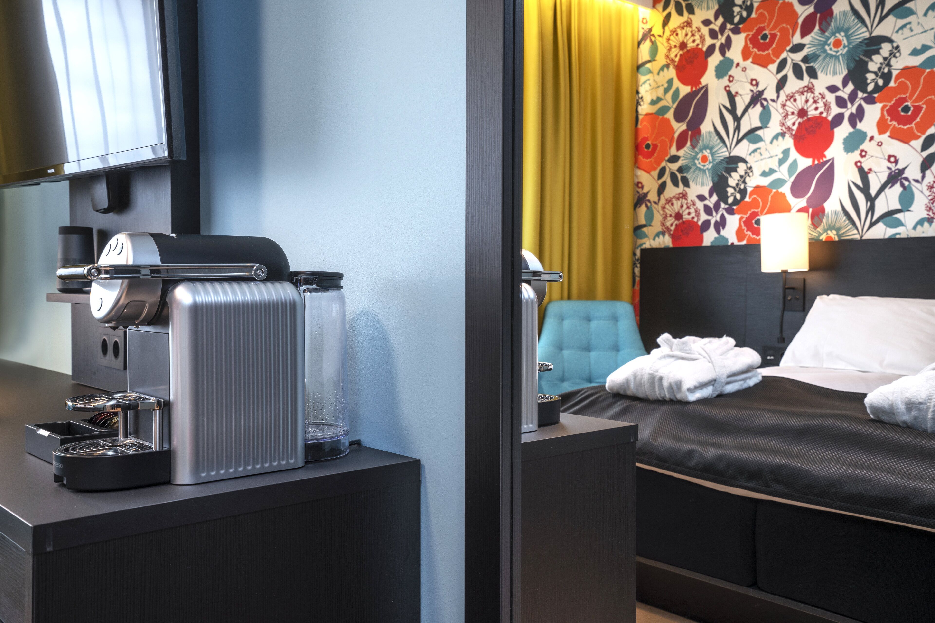 A hotel room with a bed, a bedside table with a lamp, a chair, and a coffee machine.