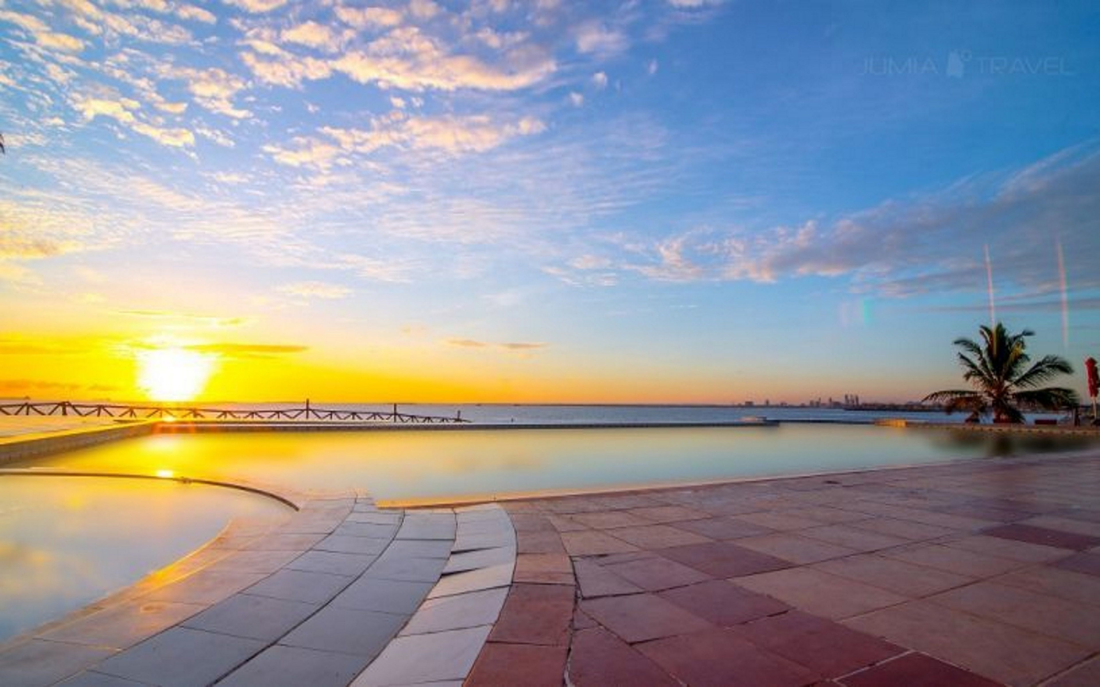 Une piscine située au bord de l’eau, offrant une vue sur le coucher de soleil, avec un palmier et un pont en arrière-plan.