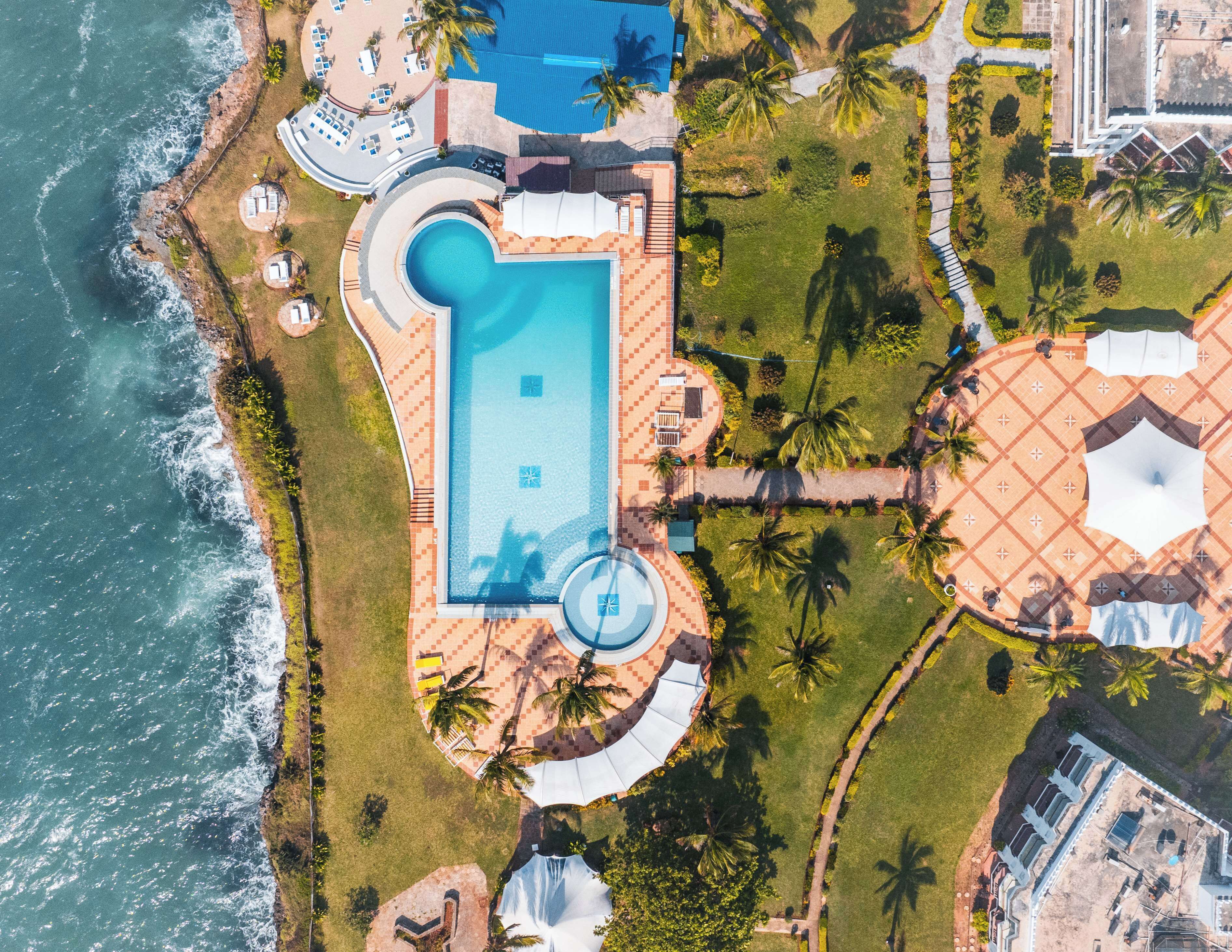Luchtbeeld van een resort met een centraal gelegen zwembad, palmbomen en een strand.