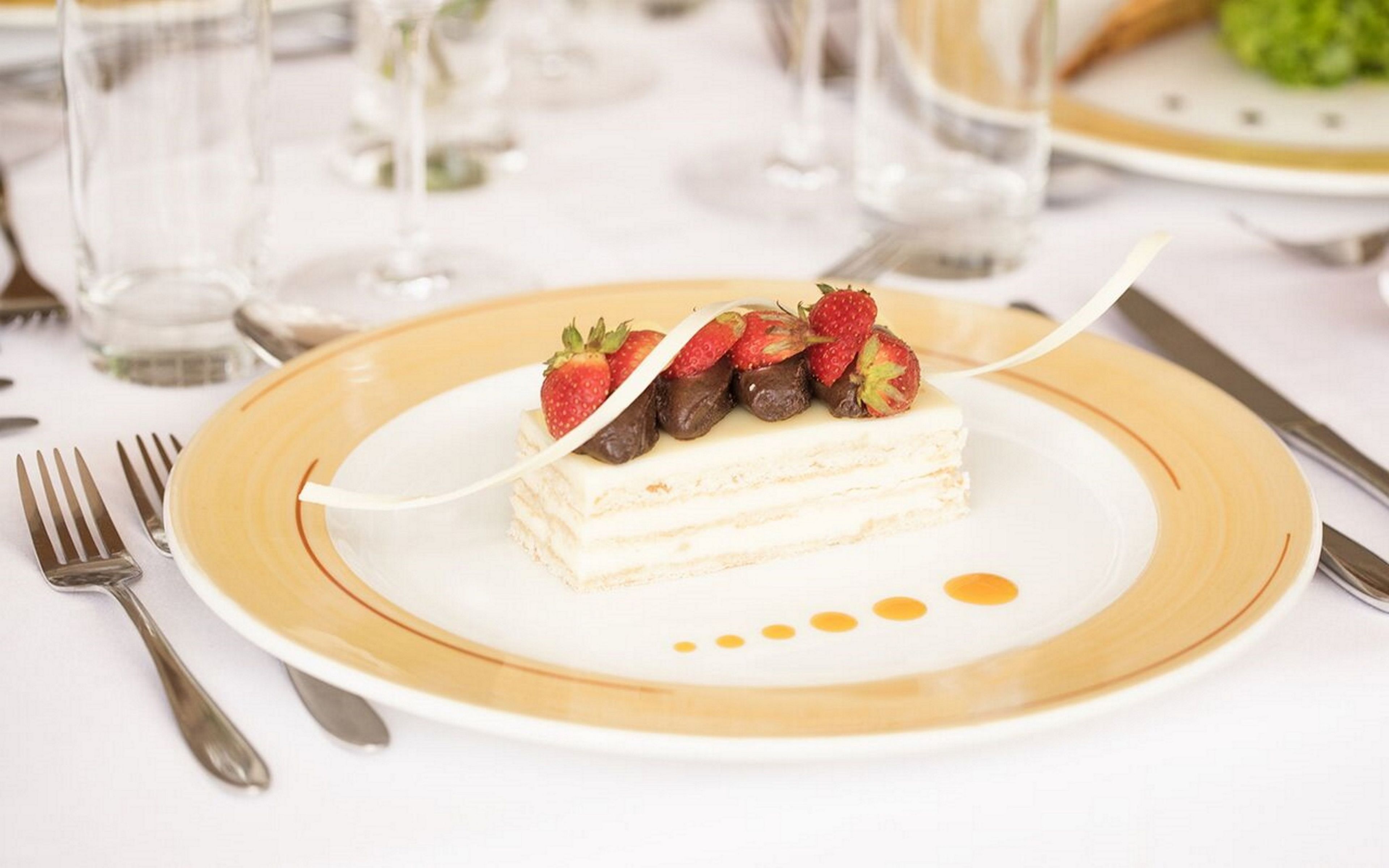 A dessert plate with a layered cake, strawberries, and chocolate pieces on a white plate with a gold rim.