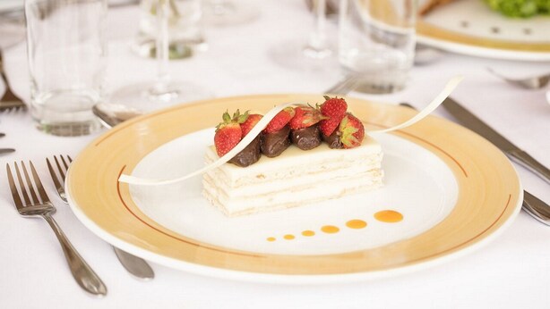 Un plato de postre con un pastel en capas, fresas y trozos de chocolate sobre un plato blanco con borde dorado.
