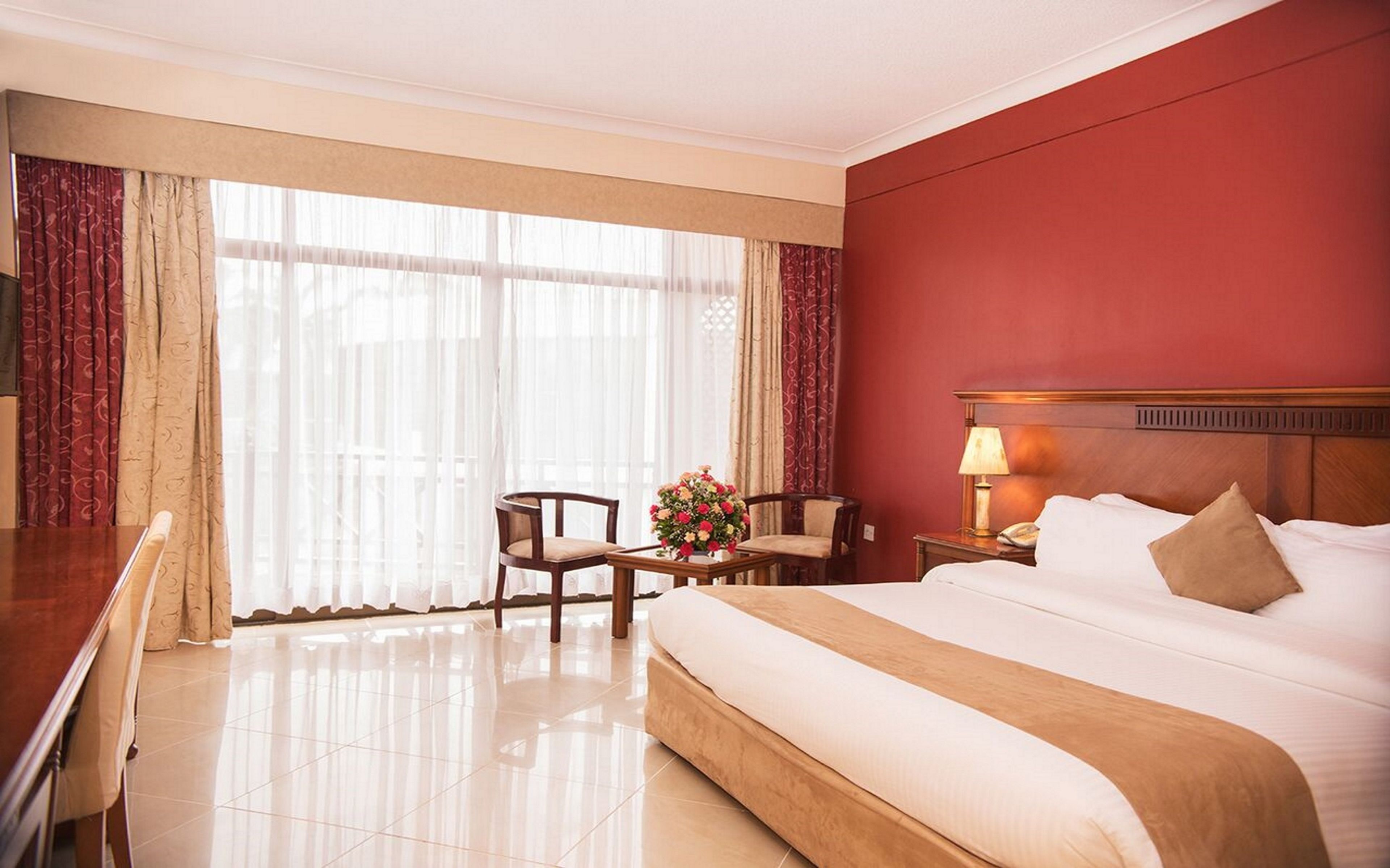 A hotel room with a large bed, a desk, two chairs, a nightstand, and a window with sheer curtains.