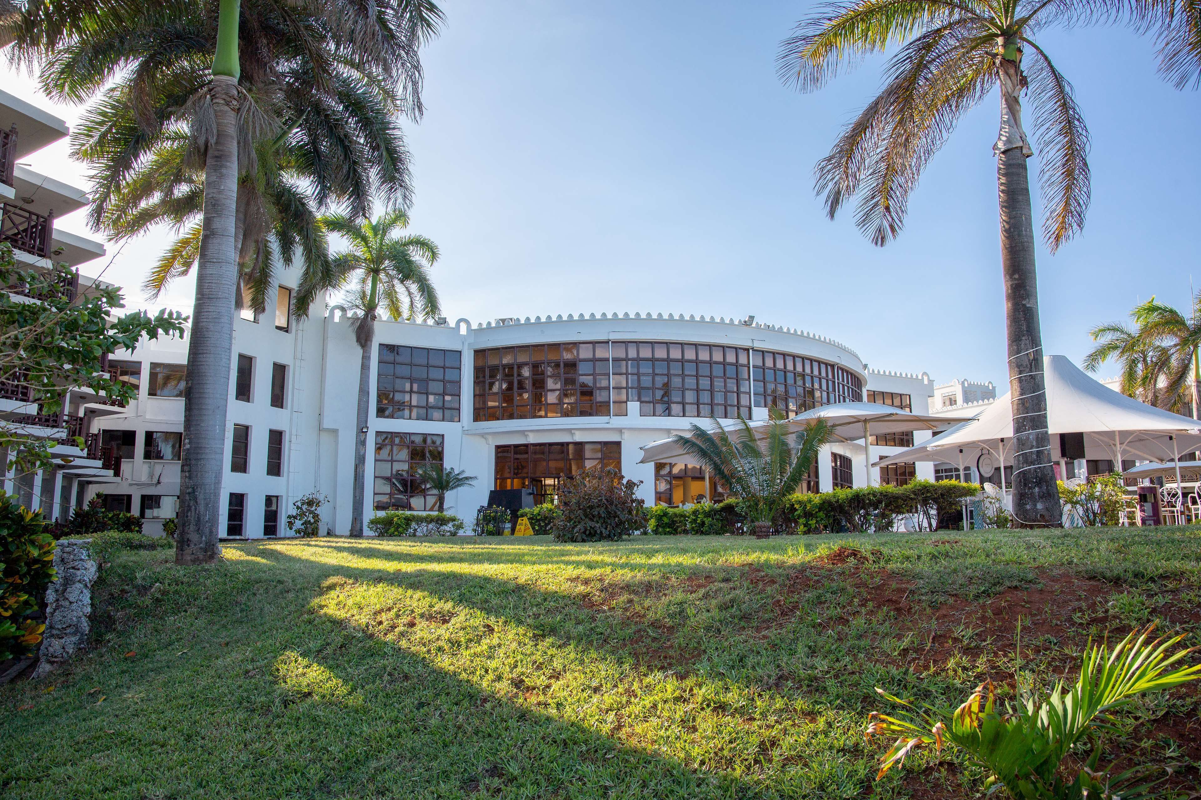 A modern hotel with a curved facade, large windows, and a well-maintained lawn with palm trees.