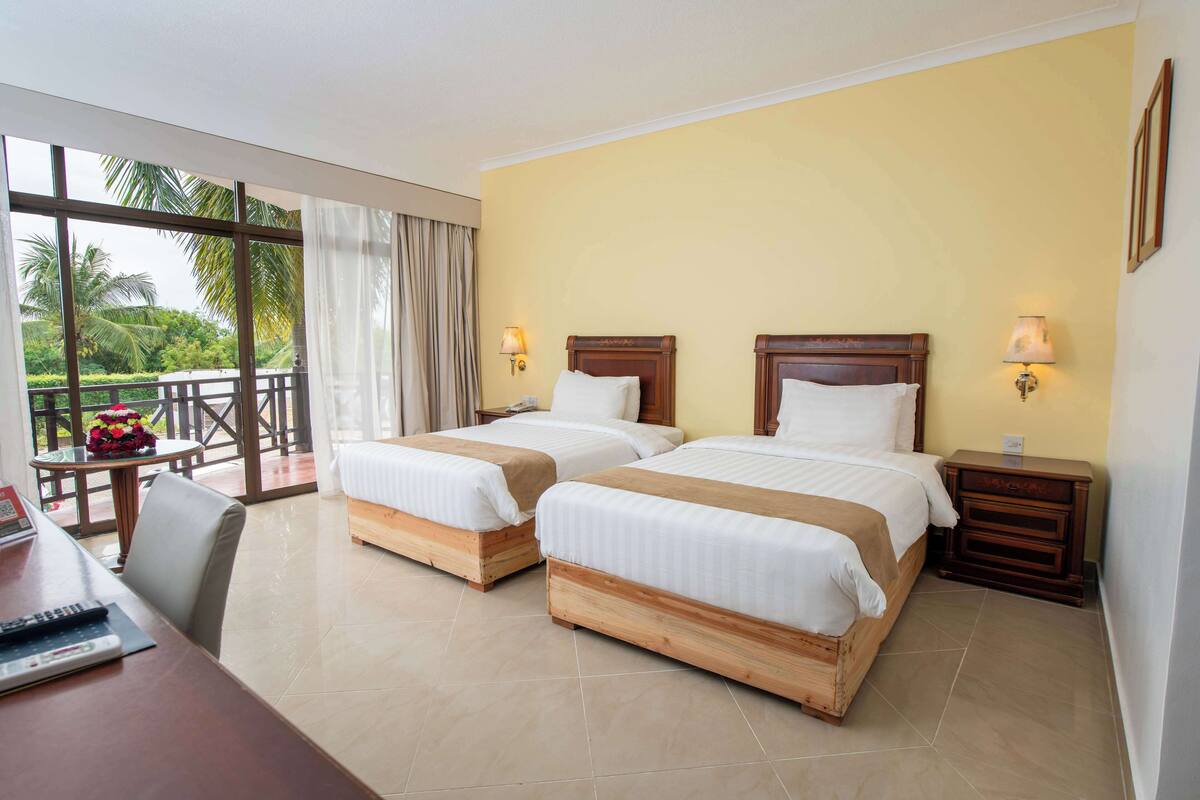 Een hotelkamer met twee bedden, een bureau en een balkon met uitzicht op palmbomen.