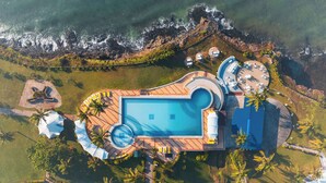 Vista aérea de un resort con piscina central, múltiples tumbonas y una zona de playa con sombrillas.