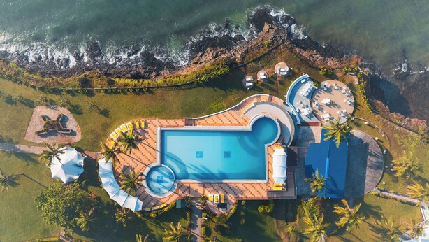 Ein Luftbild eines Resorts mit einem zentralen Schwimmbecken, mehreren Liegestühlen und einem Strandbereich mit Sonnenschirmen.