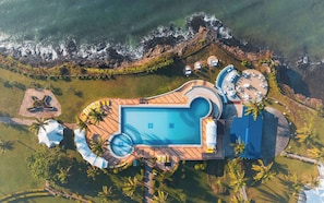Vista aérea de un resort con piscina central, múltiples tumbonas y una zona de playa con sombrillas.