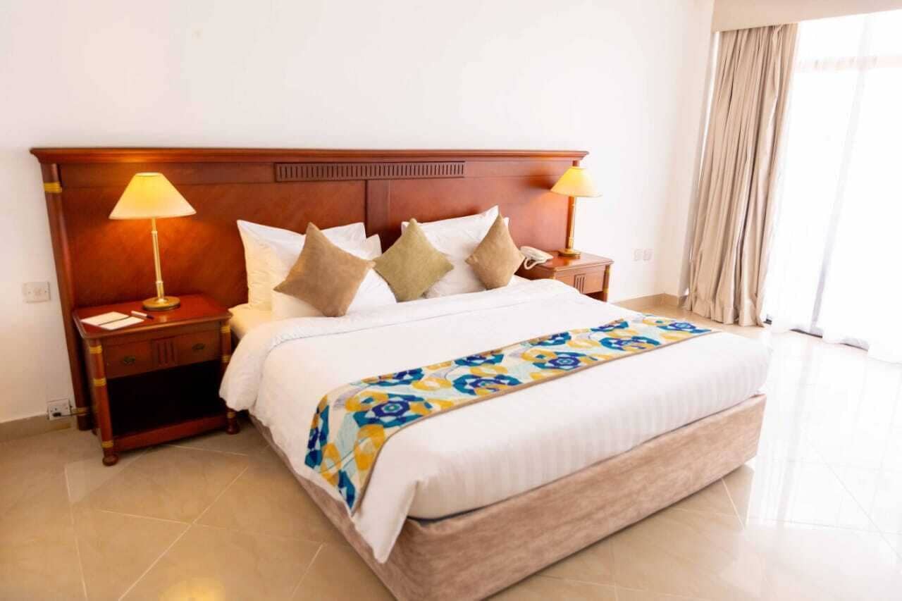 A hotel room with a large bed, two bedside lamps, a wooden headboard, a bedside table with a lamp, and a window with curtains.