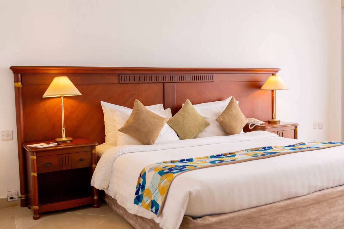 Een net opgemaakt bed met een geborduurde plaid, twee bedlampjes en een houten nachtkastje met een telefoon.