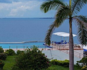 A pool area with lounge chairs and umbrellas, surrounded by greenery and overlooking a body of water.