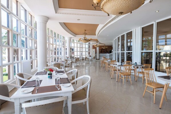 A spacious dining area with white tables and chairs, large windows, and hanging chandeliers.
