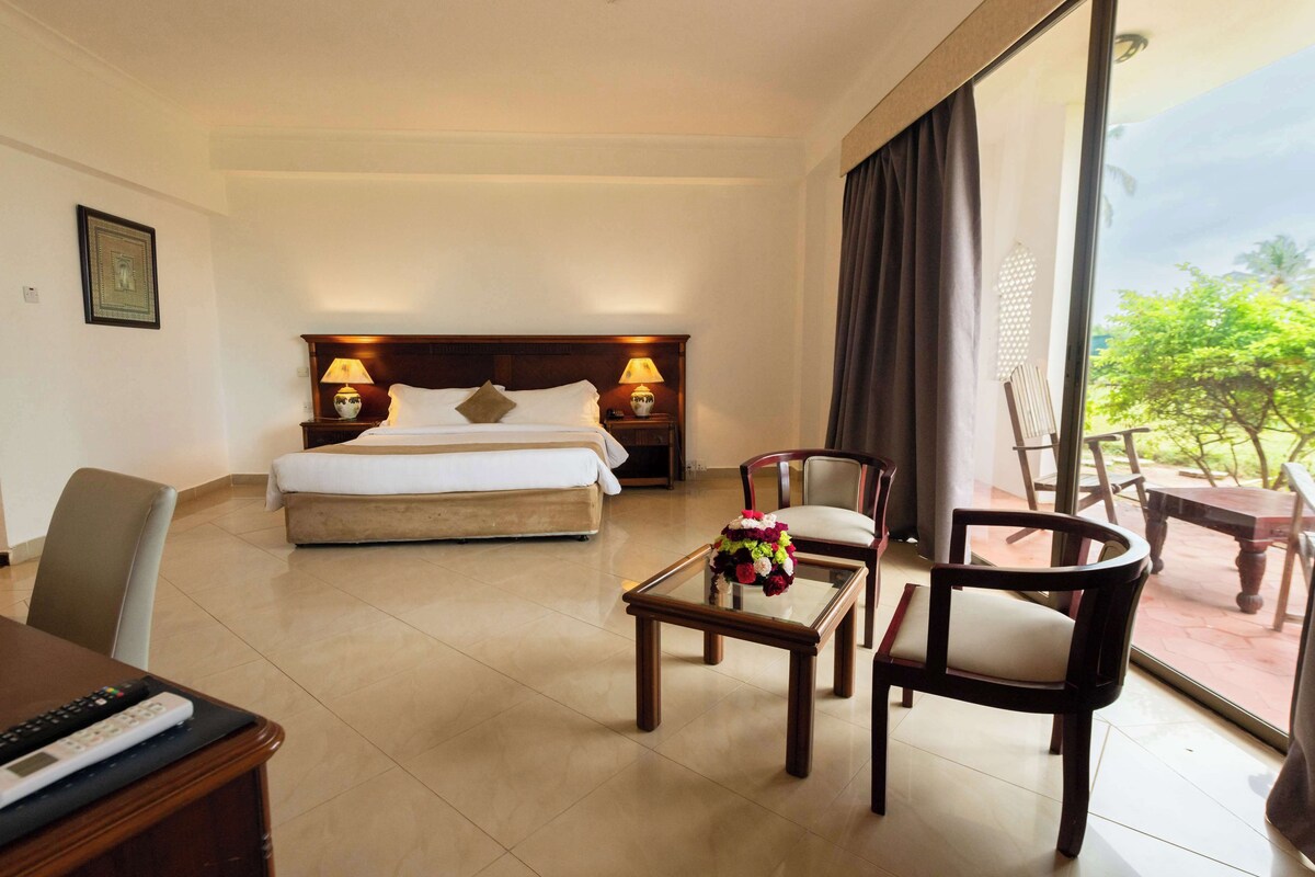 Habitación de hotel con cama, escritorio, silla, mesa con un jarrón de flores y vista al exterior.