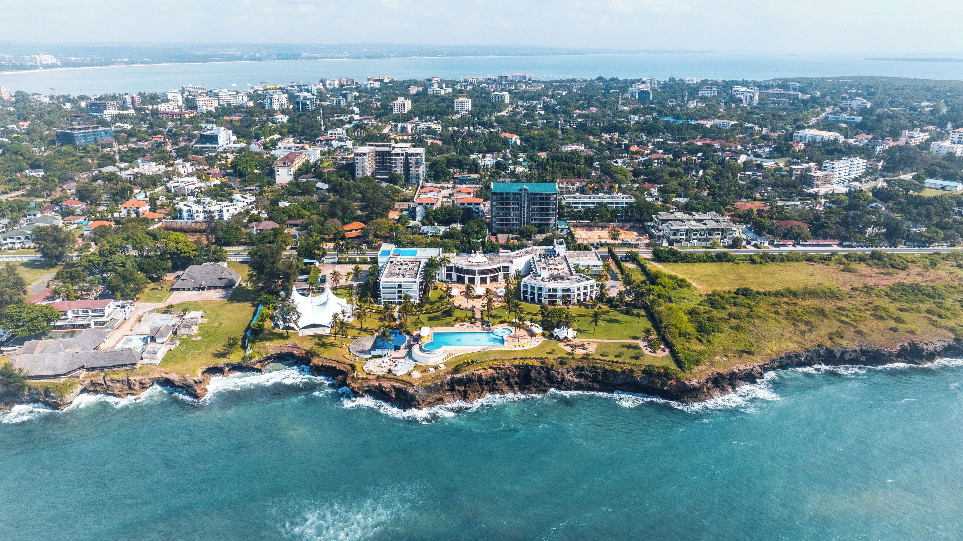 An aerial view of a coastal resort with a large pool, multiple buildings, and a rocky shoreline.