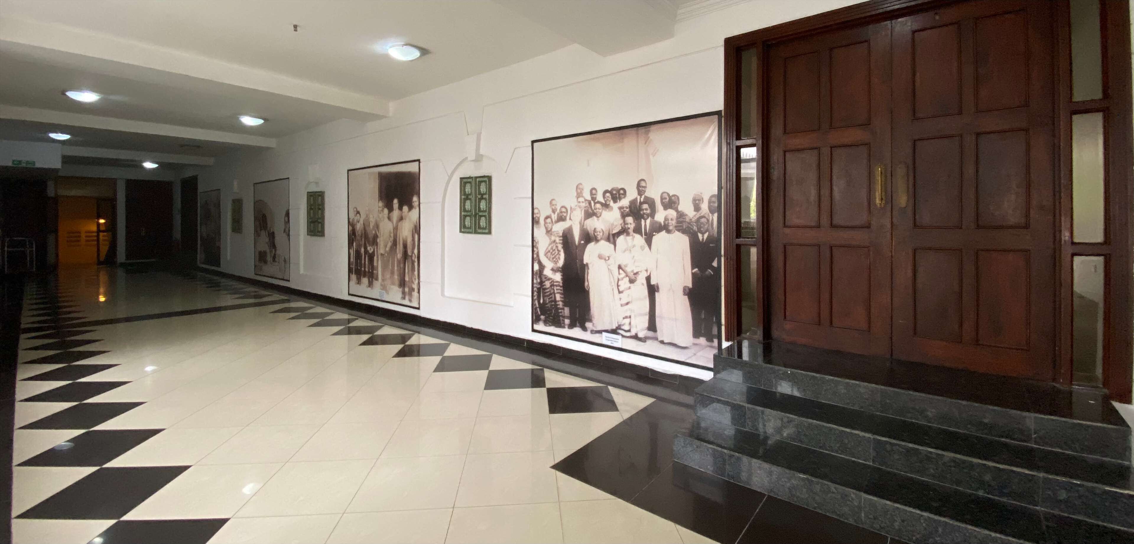 A hallway with a checkered floor, large framed photographs on the walls, and a wooden door.