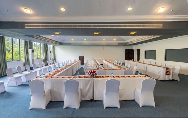 Un salón de conferencias con largas mesas, sillas blancas y un amplio ventanal con cortinas.