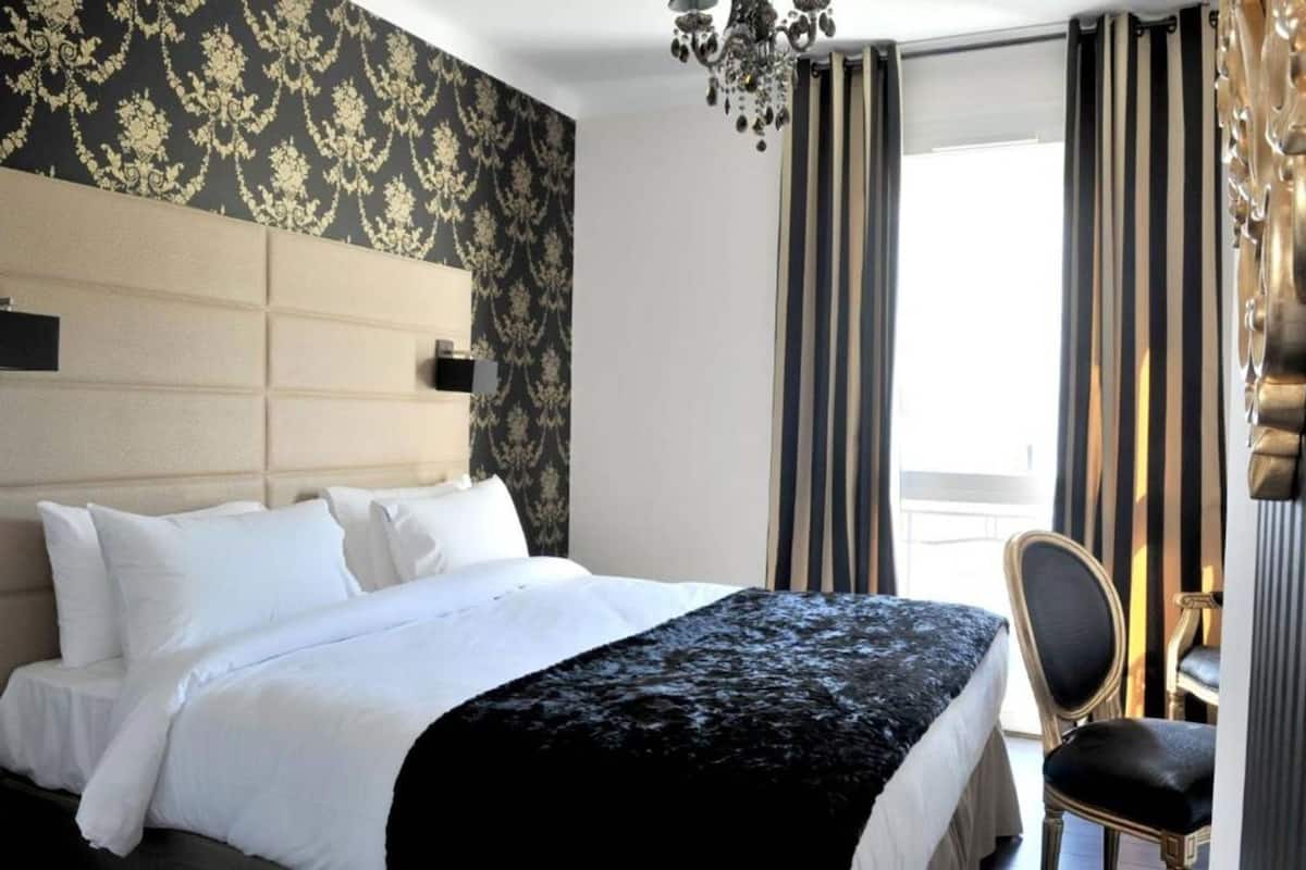 Habitación de hotel con cama, una silla, una lámpara de araña y un ventanal con cortinas.