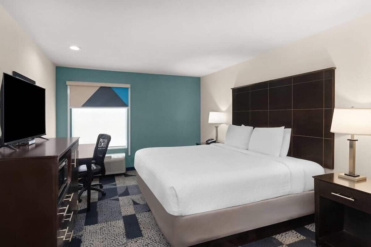 Quarto de hotel com cama, escrivaninha, cadeira, televisão e luminárias.