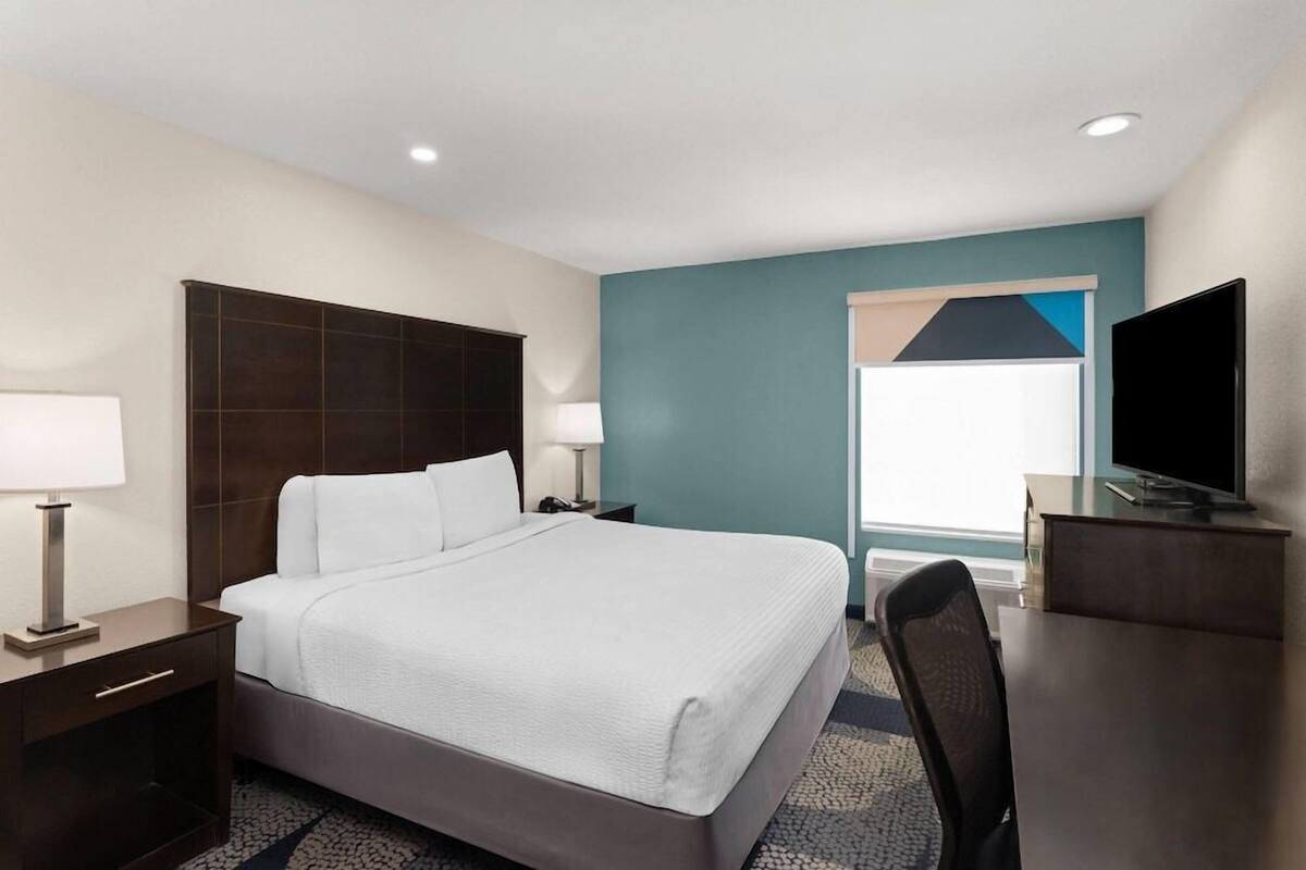 Quarto de hotel com cama, mesas de cabeceira, escrivaninha, cadeira, televisão e janela com persianas.