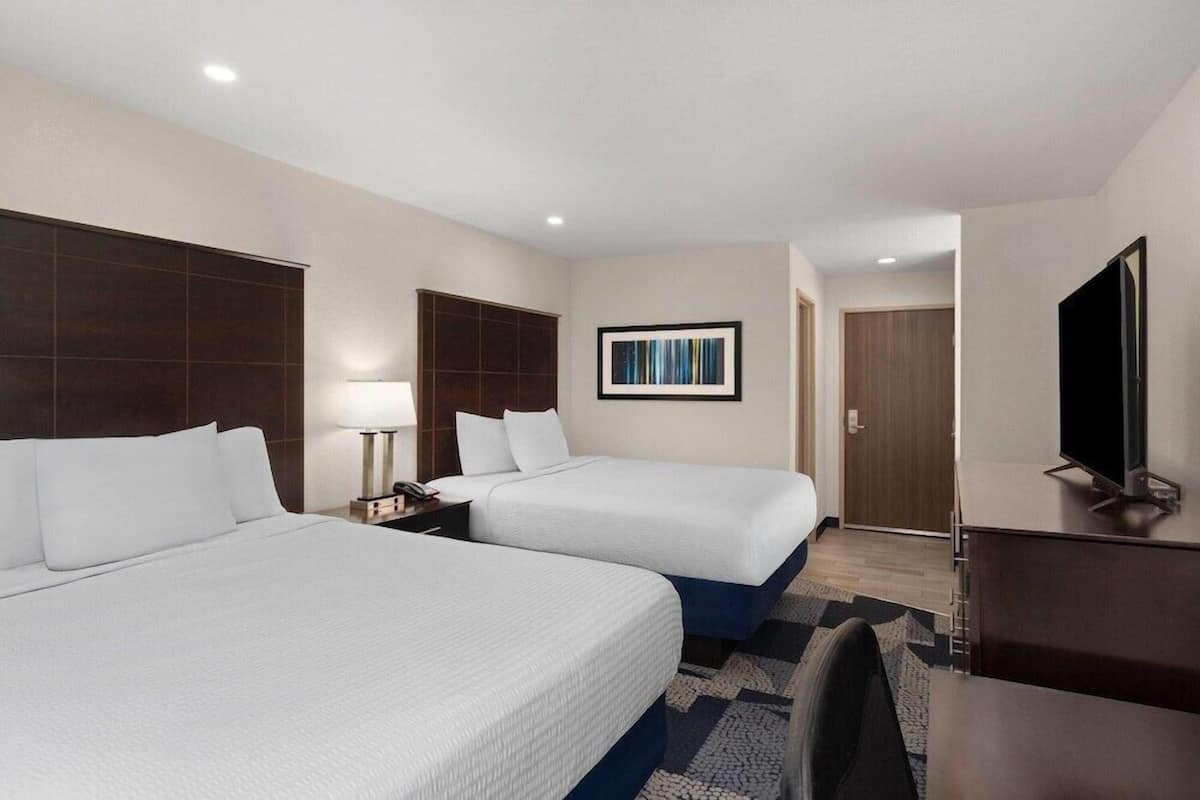Quarto de hotel com duas camas, uma escrivaninha, televisão, um quadro na parede e uma porta que dá para outro cômodo.