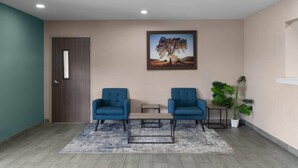 A room with two blue chairs, a small table, a framed picture of a tree, a door, and a potted plant.