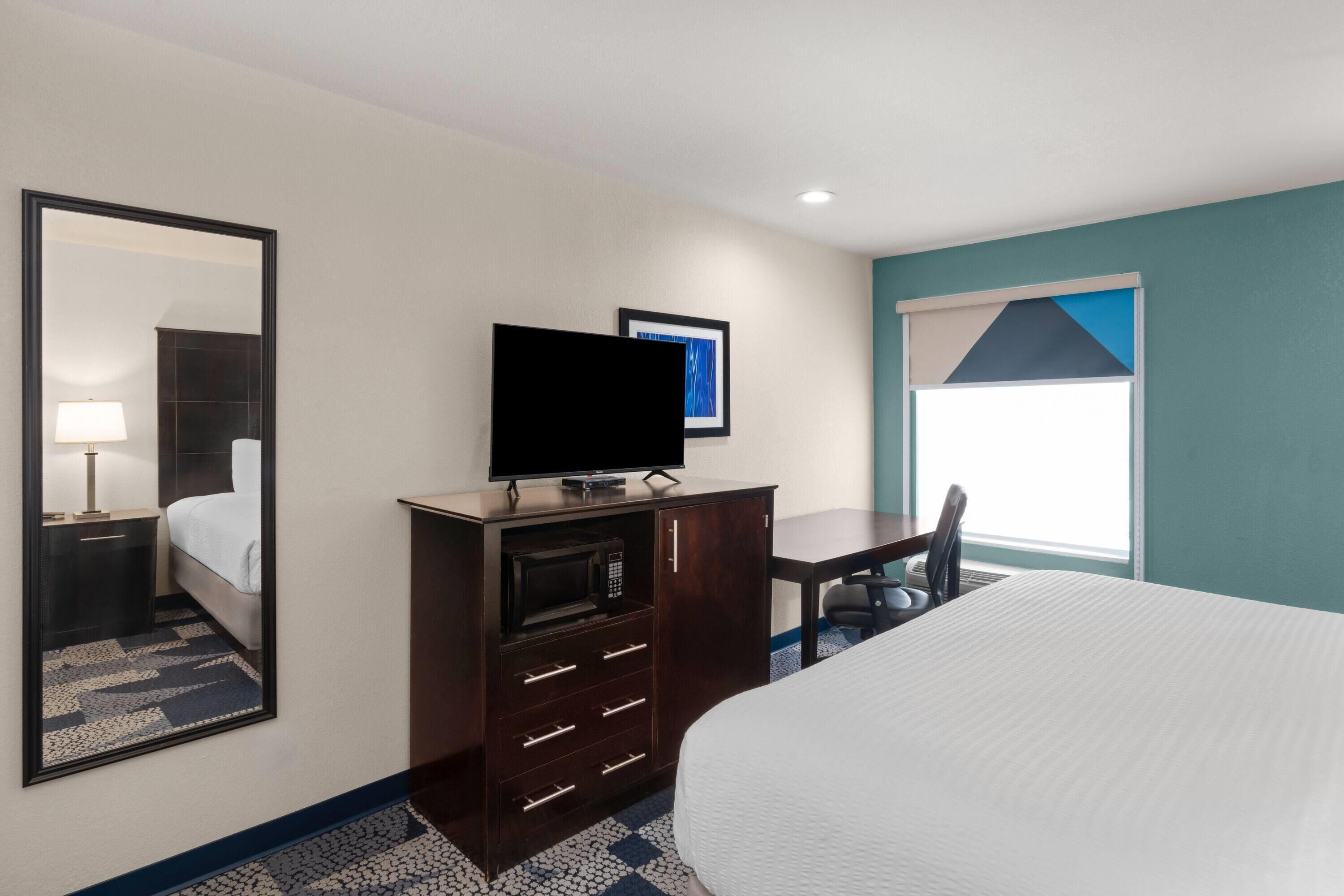 A hotel room with a bed, a TV on a wooden cabinet, a desk, and a window with blinds.