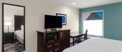 A hotel room with a bed, a TV on a wooden cabinet, a desk, and a window with blinds.