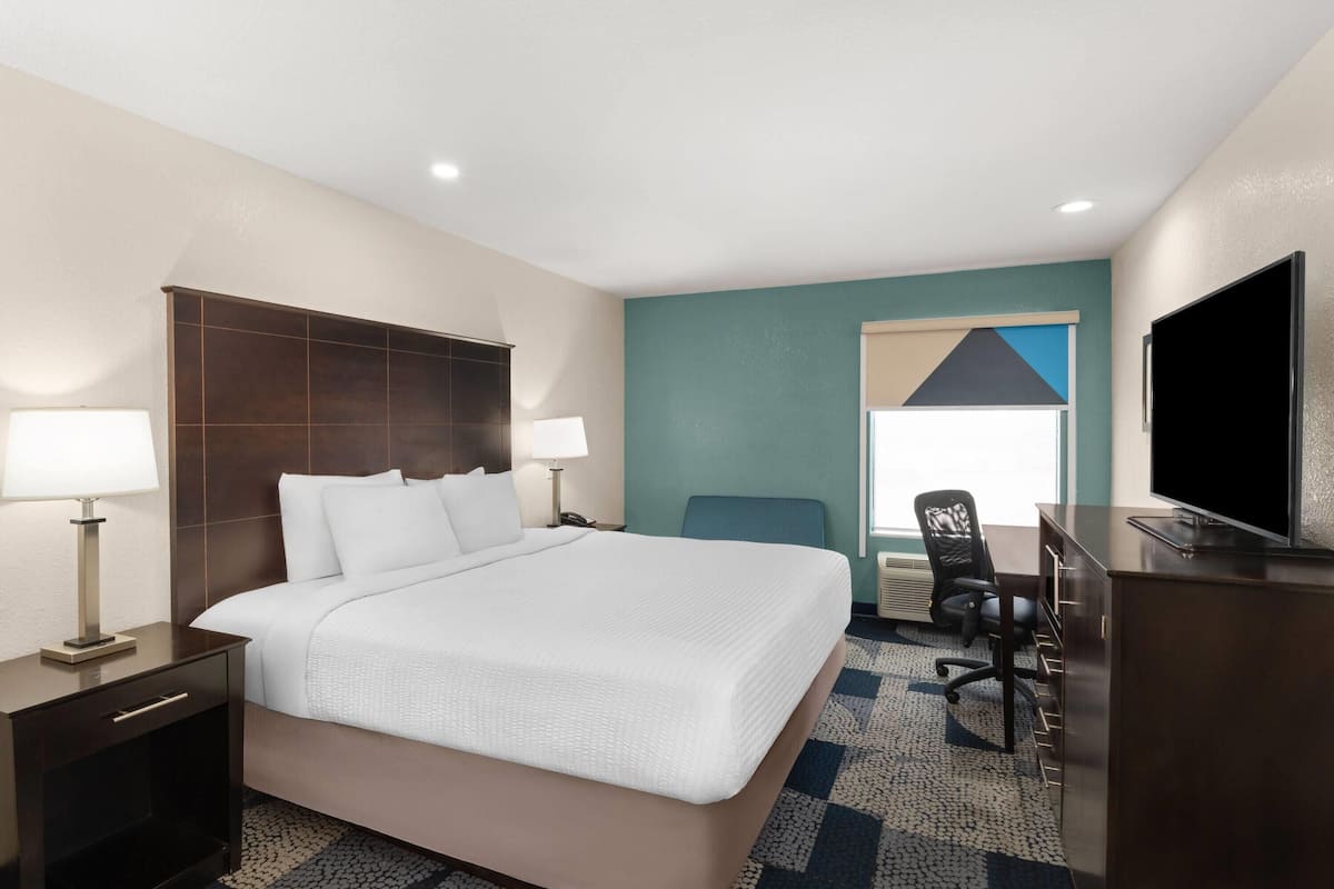 Quarto de hotel com cama, duas luminárias de cabeceira, escrivaninha com cadeira, televisão e janela com persianas.
