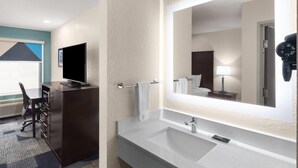 A hotel bathroom with a large mirror, a sink, a towel rack, a desk with a chair, and a television.