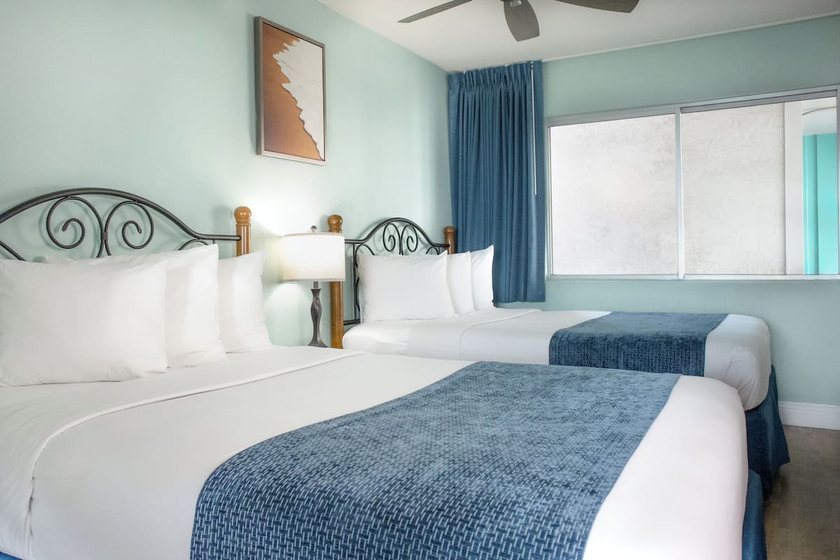 Quarto de hotel com duas camas, um ventilador de teto, janela com cortinas e um quadro na parede.