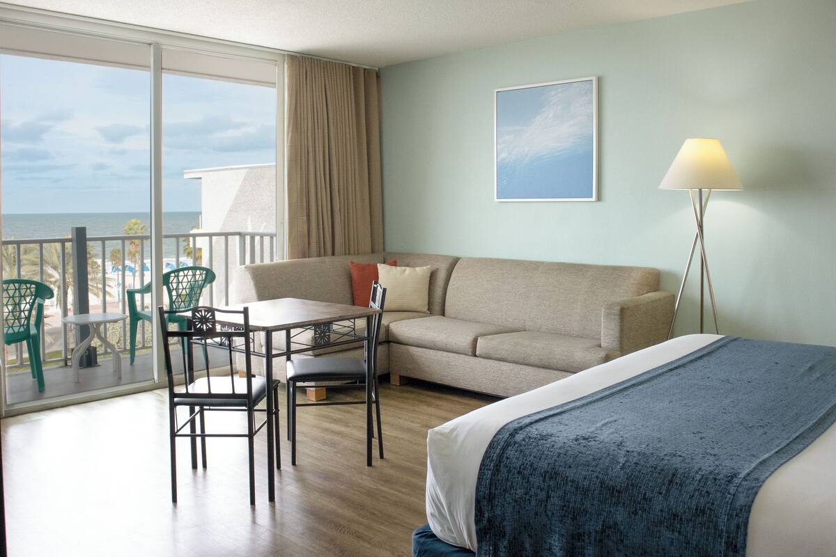 Quarto de hotel com varanda, sofá, mesa e cadeiras, e uma luminária.