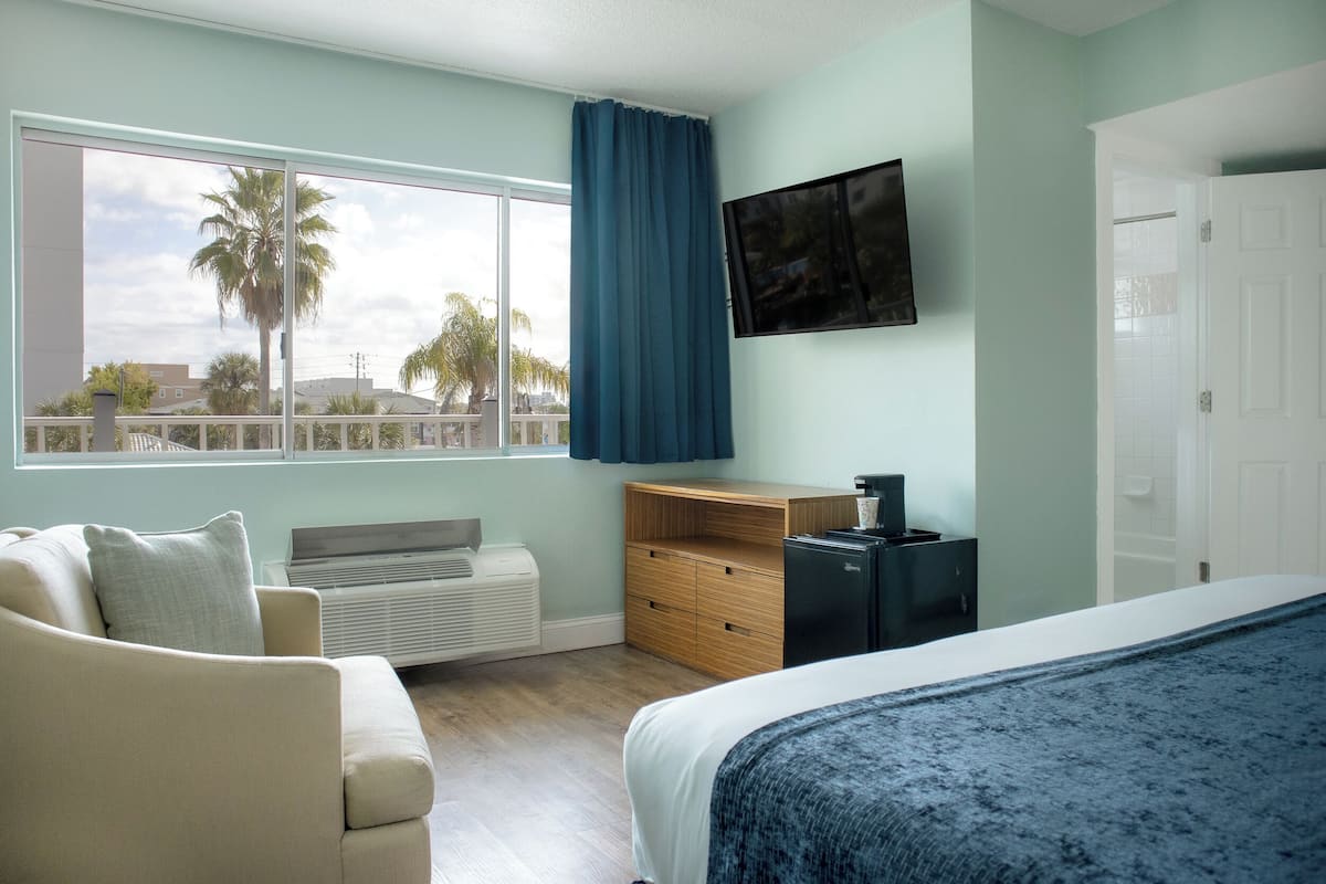 Quarto com cama, escrivaninha, cadeira, televisão e janela vista para palmeiras.