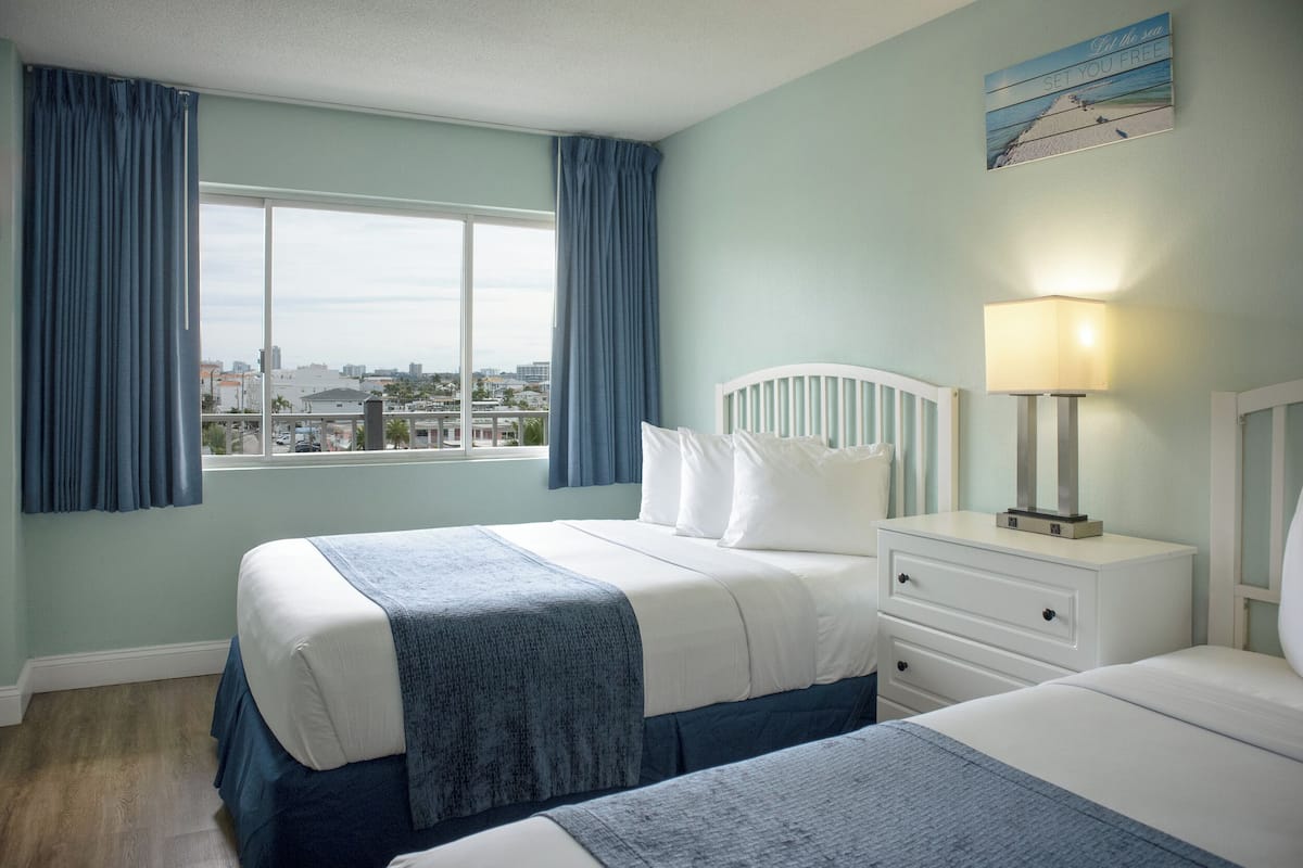 Quarto com uma cama grande, uma mesa de cabeceira com abajur e vista para a cidade pela janela.