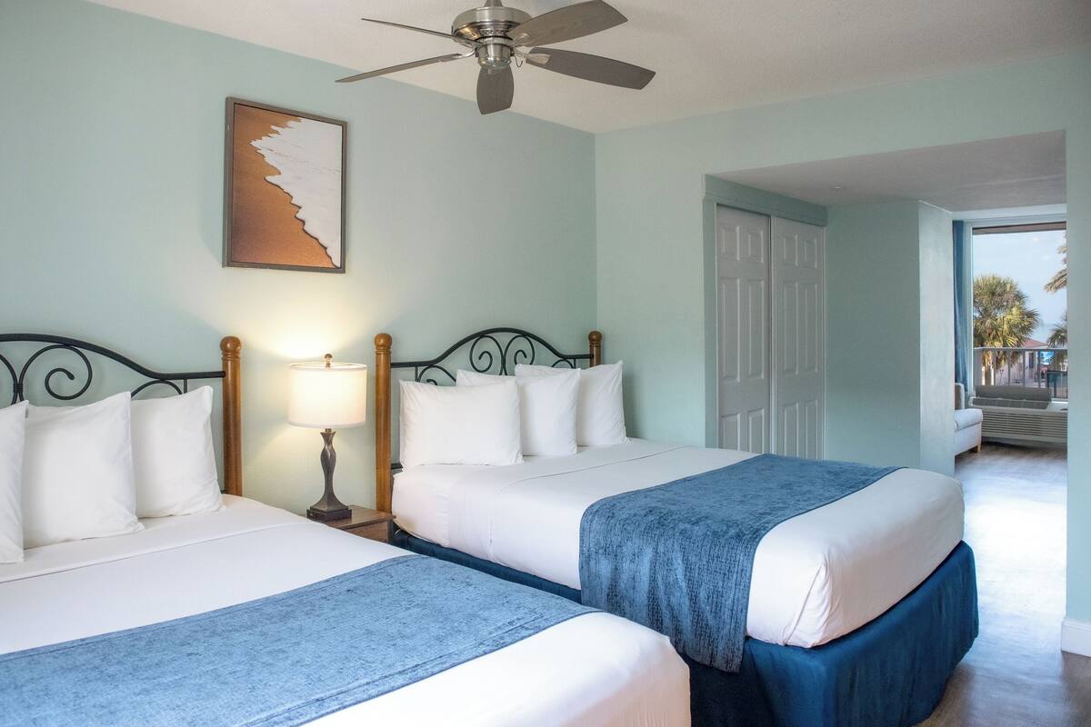 Quarto de hotel com duas camas, um ventilador de teto, uma mesa de cabeceira com luminária e um quadro na parede.