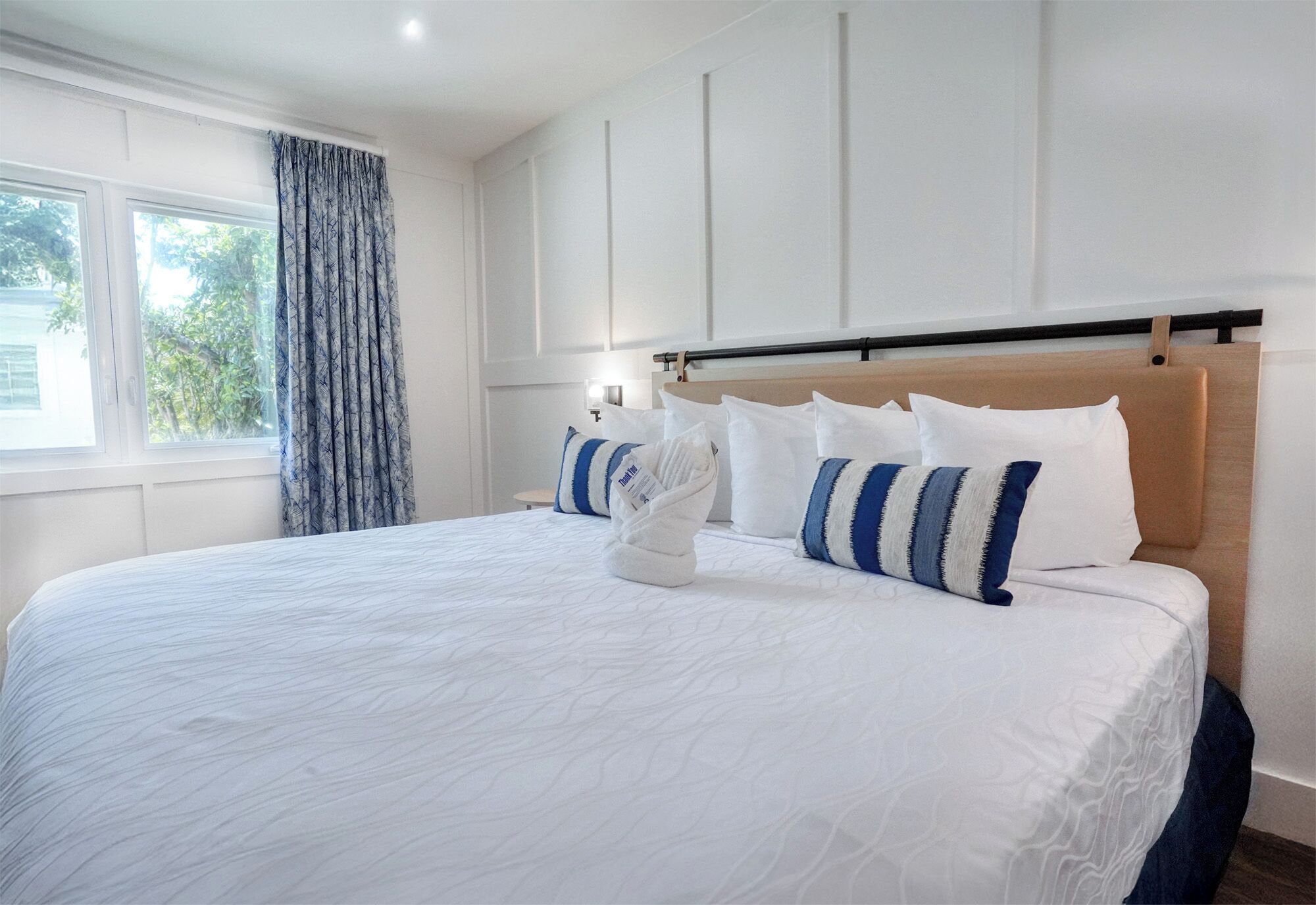 Una cama bien hecha con ropa de cama blanca y almohadas a rayas azules, un cabecero de madera y una ventana con cortina azul.