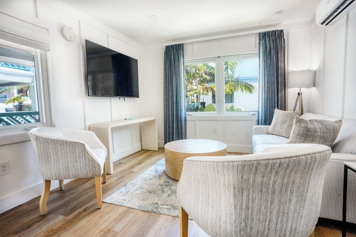 A modern living room with a flat-screen TV, a sofa, two armchairs, a coffee table, and a view of palm trees through large windows.