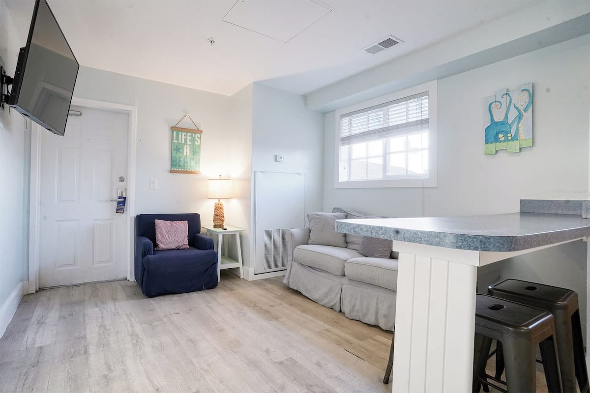 A compact living space with a kitchenette, a sofa, a chair, and a TV mounted on the wall.