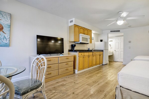 A compact kitchen with wooden cabinets, a microwave, and a refrigerator. There's a dining area with a glass table and chairs, and a bed in the room. - The Lighthouse Resort Inn & Suites (Fort Myers Beach)