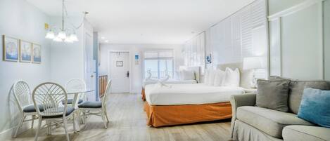 A hotel room with a bed, a sofa, a dining table with chairs, and a view of the sea through the window.